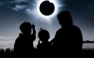 Natural phenomenon. Silhouette back view of mother and child sitting and relaxing together. Boy pointing to solar eclipse on dark sky background. Happy family spending time together. Outdoor.